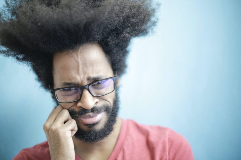 Suspicious young bearded ethnic male with creative Afro hairstyle wearing eyeglasses and pink t shirt touching cheek and looking at camera with uncertain or doubt expression. Ivy Lane Dentistry can address your tooth pain. Schedule an appointment in our San Antonio, TX location. 